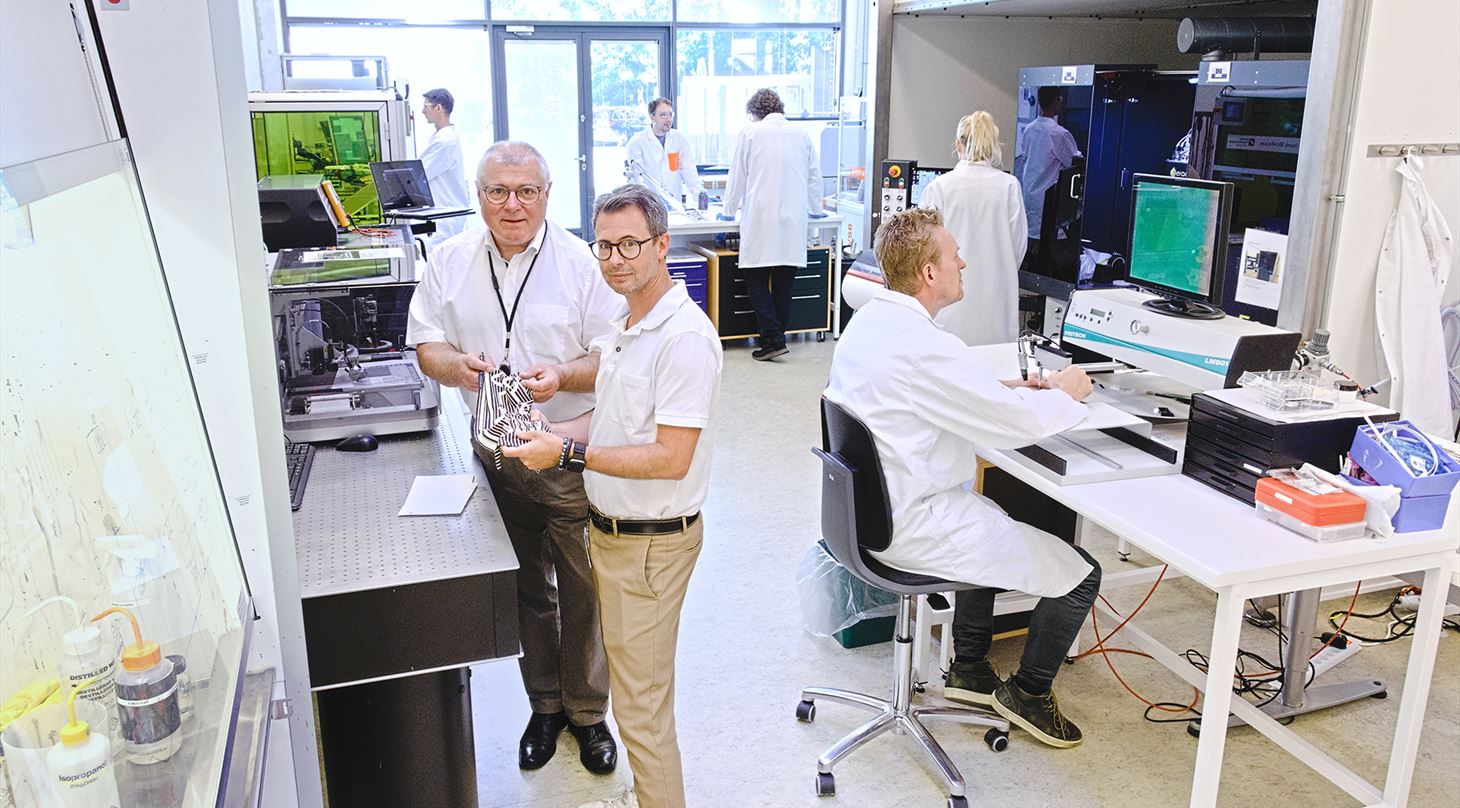 To mænd i forgrunden i laboratorie med printere